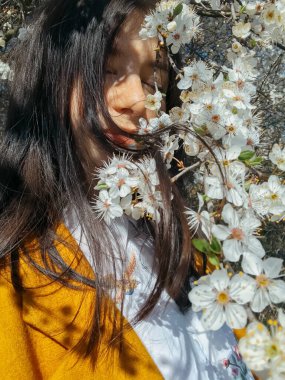 Çiçek açan kiraz dalları arasında poz veren zarif, güzel bir kadın. Şehvetli, sakin bir an. Beyaz çiçeklerle sarılmış sarı ceketli, şık, genç, esmer bir kadın.