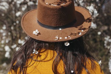 Şapkalı şık bir kadın güneşli bir günde bahar taçyapraklarıyla çiçek açan kiraz dallarında poz veriyor. Sakin bir an. Modaya uygun bir kadın beyaz çiçeklerde dinleniyor, kırsal bölgeleri keşfediyor.