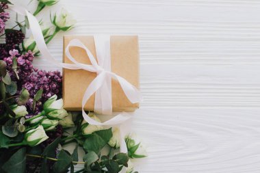 Leylak ve gül desenli şık bir hediye kutusu beyaz ahşap arka planda duruyordu, mesaj için yer vardı. Anneler günün ve kadınlar günü konseptin kutlu olsun. Kurdele ve bahar çiçekleriyle birlikte el işi.