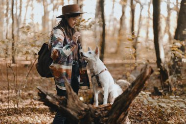 Güneşli sonbahar ormanlarında sevimli beyaz köpekleri eğiten şık bir kadın. Şirin İsviçreli çoban köpeği yavrusu muamelesiyle öğreniyor. Sonbahar ormanında sırt çantalı hippi bir kadın köpeğiyle oynuyor. Metin için boşluk