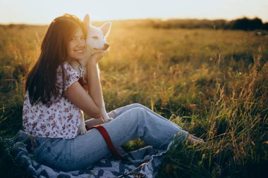 Sıcak gün batımı ışığında yaz çayırında sevimli beyaz bir köpeğe sarılan mutlu bir kadın. Sıradan genç bir bayan, sevimli bir çoban köpeğiyle rahatlıyor. Mutluluk. Evcil hayvanla yaz tatili