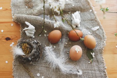 Mutlu Paskalyalar. Doğal Paskalya yumurtaları, beyaz tavşanlar, tüy, yuva ve kiraz çiçekli dallar masanın üzerinde kırsal keten peçeteler. Üst Manzara. Mesaj için yer var. Çevre dostu dekor
