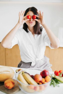 Genç, mutlu bir kadın, elinde vişneli domatesle modern mutfakta taze meyve ve sebzelerle gülümsüyor. Sağlıklı beslenme ve ev yemeği kavramı. Eğleniyor ve salata yapıyor.