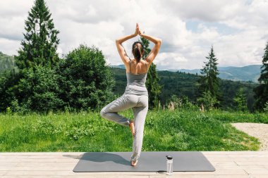 Yoga yapan ve güneşli dağların arka planında poz veren zinde bir kadın. Açık hava antrenmanı ve antrenman. Sağlıklı bir yaşam tarzı. Spor Dişi, ağaçların arasındaki tahta terasta yoga yapıyor.
