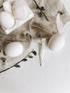 Natural easter eggs, soft feathers, pussy willow branches on rustic cloth on white aged table. Flat lay. Simple stylish rural Easter still life. Modern aesthetics, pastel beige and grey colors