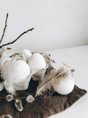 Natural easter eggs, feathers, pussy willow branches on rustic cloth on white aged table. Stylish rural Easter still life. Modern simple aesthetics,  white and grey colors