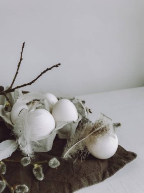 Stylish rural Easter still life. Natural easter eggs, feathers, pussy willow branches on rustic cloth on white aged table. Modern simple aesthetics, white and grey colors