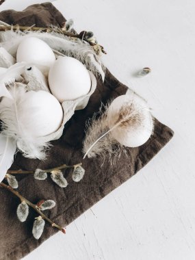 Stylish rural Easter still life. Natural easter eggs, feathers, pussy willow branches on rustic cloth on white aged table. Modern simple aesthetics, white and grey colors