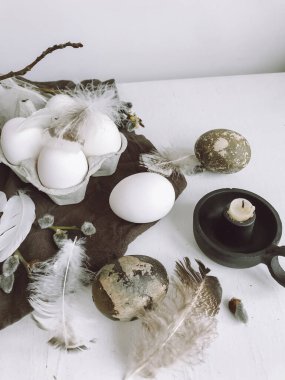 Simple stylish Easter still life. Natural easter eggs, soft feathers, pussy willow branches, candle on white rustic aged table. Modern Natural easter aesthetic, white and grey colors