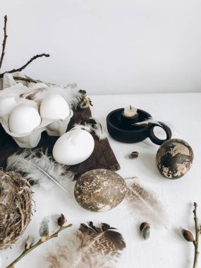 Natural easter eggs, feathers, pussy willow branches, nest and candle on rustic cloth on white aged table. Stylish rural Easter still life. Modern simple aesthetics,  white and grey colors