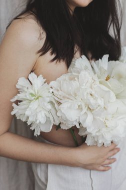 Beyaz şakayık çiçekleri olan zarif, şehvetli bir kadın. Bej kumaş arka planda pantolondan çıkan şakayık desenli buketi olan güzel bir kadın. Açan vücut pozitiftir ve çiçek kokar. Kişisel bakım