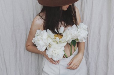 Şapkalı şık bir kadın pantolonundan çıkan beyaz şakayık çiçekleriyle poz veriyor. Bej kumaş arka planında şakayık demeti olan şehvetli genç bir kadın. Çiçek kokusu ve kişisel bakım konsepti