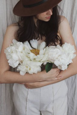 Şapkalı şık bir kadın pantolonundan çıkan beyaz şakayık çiçekleriyle poz veriyor. Bej kumaş arka planında şakayık demeti olan şehvetli genç bir kadın. Çiçek kokusu ve kişisel bakım konsepti