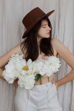Şapkalı şık bir kadın pantolonundan çıkan beyaz şakayık çiçekleriyle poz veriyor. Bej kumaş arka planında şakayık demeti olan şehvetli genç bir kadın. Çiçek kokusu ve kişisel bakım konsepti