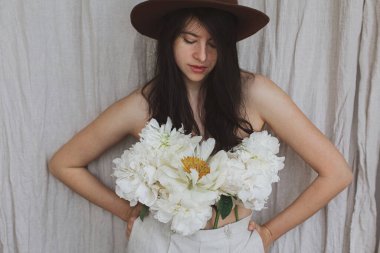 Şapkalı şık bir kadın pantolonundan çıkan beyaz şakayık çiçekleriyle poz veriyor. Bej kumaş arka planında şakayık demeti olan şehvetli genç bir kadın. Çiçek kokusu ve kişisel bakım konsepti