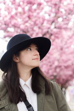 Pembe sakura ağaçlarının arka planında duran şapkalı güzel bir kadın. Genç ve şık bir bayan şehrin baharında eğleniyor.