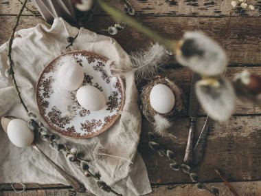 Rural Easter still life. Natural easter eggs, feathers, vintage plate and cutlery, napkin, pussy willow branches on aged wooden table, flat lay. Happy Easter. Stylish rustic Easter table setting