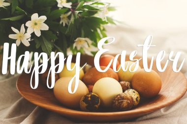 Modern Paskalya yumurtaları üzerine mutlu paskalya metinleri ahşap kasede bahar çiçekleri kırsal çarşaf bezinde el yazısıyla yazılmış. Güzel, şık bir tebrik kartı. Mevsimler selamlar