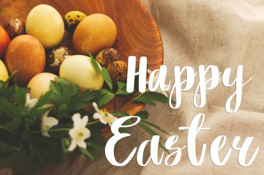 Mutlu Paskalya tebrik kartları. Modern paskalya yumurtalarının üzerine el yazısıyla yazılmış mutlu paskalya yazıları. Üzerinde bahar çiçekleri olan ahşap bir kâse. Güzel kartpostallar, mevsimler selamlar