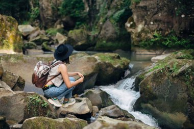 Sırt çantalı şık bir kadın gezgin dağlarda nehirde oturuyor. Genç bir kadın, günlük giysiler içinde ve şapkalı nehirde kayaların üzerinde dinleniyor. Seyahat ve seyahat tutkusu. Ormanı keşfediyorum.