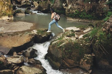 Sırt çantalı şık bir kadın gezgin dağlarda nehir taşlarında yürüyor. Giyim kuşam ve şapkayla nehrin karşısına atlayan genç bir kadın. Seyahat ve seyahat tutkusu. Yürüyüş ve ormanı keşfetmek.