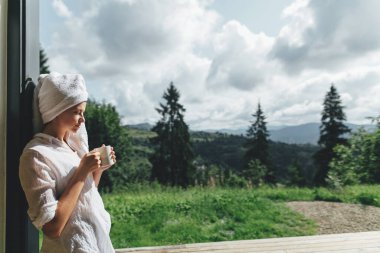 Güzel genç bayan, dağ tepelerinin arkasında bir fincan kahveyle güneşli bir sabahın tadını çıkarıyor. Sakin bir an, mesaj için yer. Güzel, rahat, beyaz pijama ve havlu giymiş kahve içen bir kadın.
