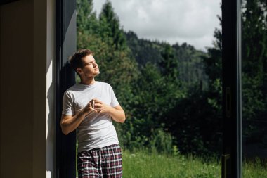 Yakışıklı adam, dağ tepelerinin arka planında güneşli bir sabahta sıcak kahvenin tadını çıkarıyor. Sakin bir an. Pijamalı genç adam verandada bir fincan kahveyle rahatlıyor. Metin için boşluk