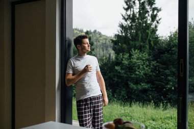 Yakışıklı adam, dağ tepelerinin arka planında güneşli bir sabahta sıcak kahvenin tadını çıkarıyor. Sakin bir an. Pijamalı genç adam verandada bir fincan kahveyle rahatlıyor. Metin için boşluk