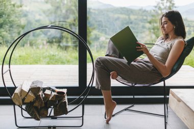 Güzel stil sahibi bir kadın şöminedeki sandalyede kitap okuyor ve dağ tepelerinde yakacak odunları var. Gündelik giysiler içinde genç bir kadın modern dağ evinde dinleniyor ve pencereden manzarası harika.