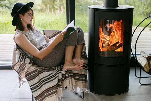 Beautiful stylish woman reading book on chair at warm burning fireplace on background of big window. Young female in casual clothes relaxing in modern chalet, cozy vacation. Leisure time