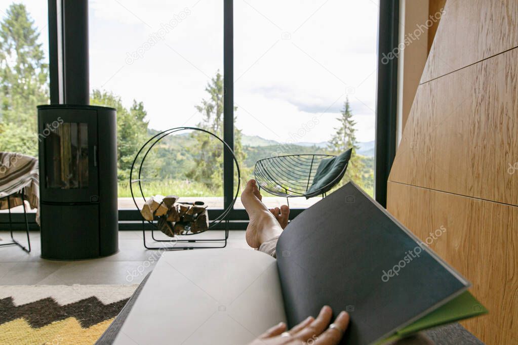 Beautiful female feet and book on background of  fireplace and big window in modern chalet. Young woman barefoot relaxing with book in comfortable home, cozy vacation. Leisure time