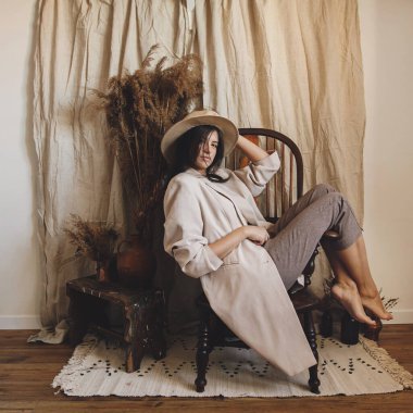 Bej takım elbiseli şık bir kadın ve bohem stil odasında ahşap koltukta poz veren bir şapka. Kurumuş çimenler ve vazoda çiçekler. Genç ve çekici bir kadın atmosferik boho iç mekanında oturuyor.