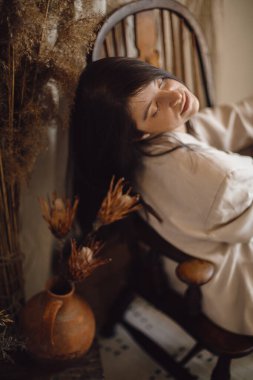 Kurumuş çimenler ve vazoda çiçeklerle bohem tarzı odada bej takım elbiseli şık bir kadın ahşap sandalyeye poz veriyor. Genç ve çekici bir kadın atmosferik boho iç mekanında oturuyor.