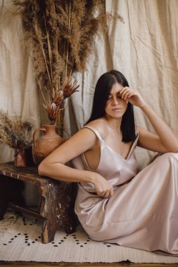 İpek elbiseli şık bir kadın bej kumaş arka planında kuru çimenler ve çiçeklerle bohem tarzı poz veriyor. Genç ve çekici bir kadın atmosferik boho iç mekanında oturuyor.