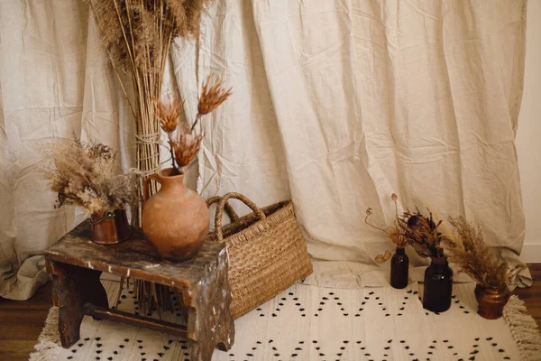 Decor details of atmospheric bohemian style interior room. Dry protea flowers in clay vase, pampas grass, wicker basket, rug on background of beige cloth. Boho decor for wedding or in studio