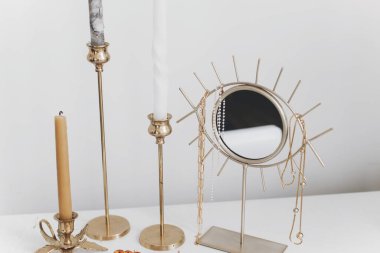 Altın kolyesi ve beyaz masasında klasik mumları olan modern altın boho aynası. Şık altın aksesuarlar, zincir kolye ve inciler. Boho mücevherleri ve detayları.