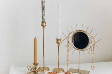 Altın kolyesi ve beyaz masasında klasik mumları olan modern altın boho aynası. Şık altın aksesuarlar, zincir kolye ve inciler. Boho mücevherleri ve detayları.