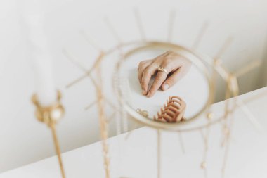 Modern altın yüzüklü ve küpeli kadın eli beyaz masadaki klasik mumlarla boho aynasına yansıyor. Şık altın yüzük, kolye, saç tokaları. Boho mücevherleri.