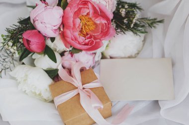 Güzel bir şakayık buketi, tebrik kartı ve kırsal sandalyede yumuşak beyaz kumaş üzerinde hediye kutusu. Pembe ve beyaz şakayık çiçekleri, hediye, kartpostal. Anneler Günün kutlu olsun. Düğün davetiyesi