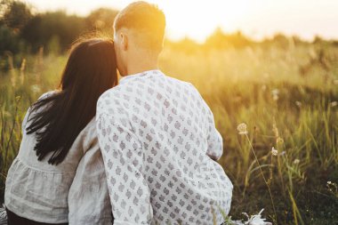 Güzel bir çift gün batımının tadını çıkarıyor, yaz otlağında çimlerin arasında sıcak güneşli bir ışıkta battaniyede dinleniyorlar. Tatil ve piknik. Genç boho ailesi kucaklıyor, atmosferik güzel bir an.