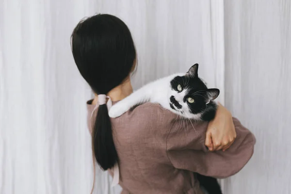 穿着亚麻布衣服的时髦女人 背着可爱的猫 衬托着瓷布 简单而缓慢的生活年轻的女性穿着粗俗的衣服抱着可爱的黑白猫咪 可爱的一刻 — 图库照片