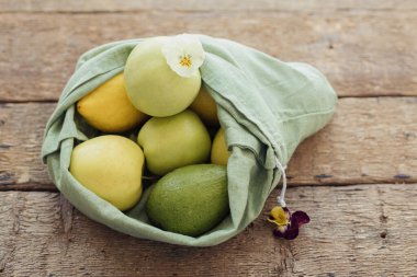 Sıfır atık alışveriş kavramı. Taze elmalar, avokado, eko pamuklu limonlar ve kır ağacından yapılmış masa çiçekleri. Yeşil poşette organik meyve ve sebzeler. Çevre dostu plastikler.
