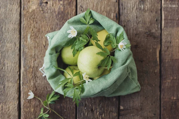 Kırsal ahşap masada eko pamuk çuvalında taze elmalar. Sıfır atık alışveriş kavramı. Plastiği yasakla. Yeşil poşette organik elmalar ve çiçekler, üst manzara. Çevre sürdürülebilir yaşam biçimi