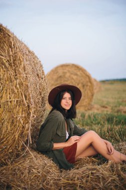 Yaz akşamları saman balyasında oturan şapkalı güzel bir kadın. Mutlu genç bayan samanlıkta dinleniyor, yaz tatilinde kırsalda. Sükunet, kırsal yaşam