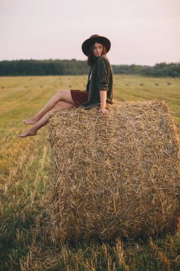 Güzel, mutlu bir kadın yaz samanlığında gün batımında samanlıkta oturuyor. Kırsalda saman balyasında rahatlayan genç ve mutlu bir dişi. Atmosferik sükunet anı