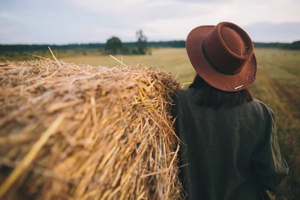 夏の夜のフィールド バックビューで干し草の山に立ってわらで帽子のスタイリッシュな女性 田舎の静かな瞬間 若いです女性休息に干し草ベール — ストック写真
