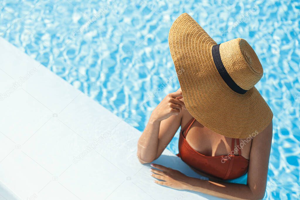 Beautiful woman in hat relaxing in pool water, enjoying summer vacation at tropical resort. Slim young female sunbathing at swimming pool edge, view above. Holidays and travel. Space for text