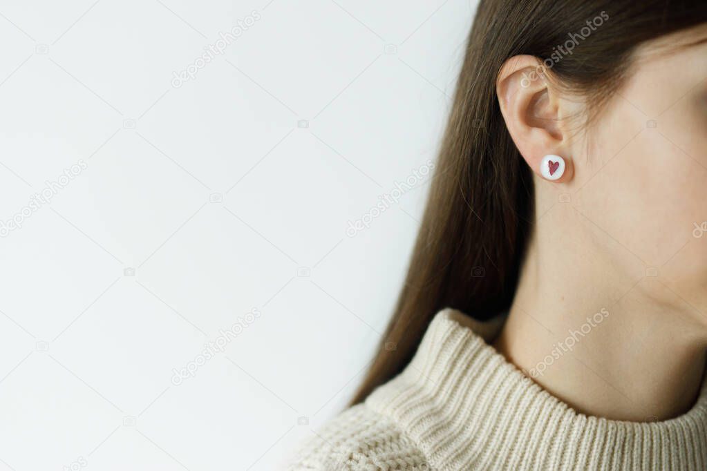 Beautiful stylish woman with cute earring with red heart, cropped view. Fashionable female in sweater with unusual fused glass accessory. Beauty and care. Valentine gift. Copy space