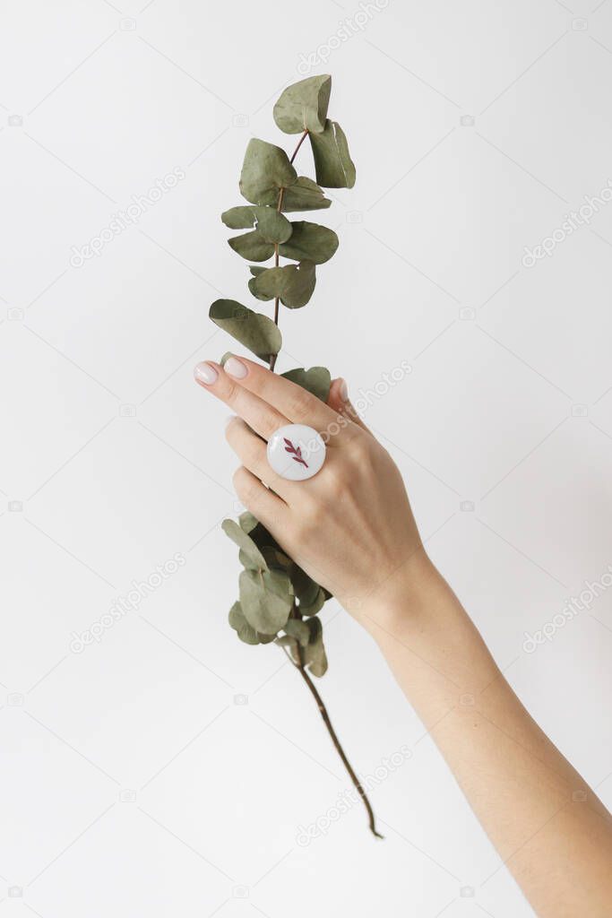 Stylish modern white round ring on beautiful hand with eucalyptus branch on white background. Unusual fashionable fused glass ring on female hand with white manicure. Eco and care concept