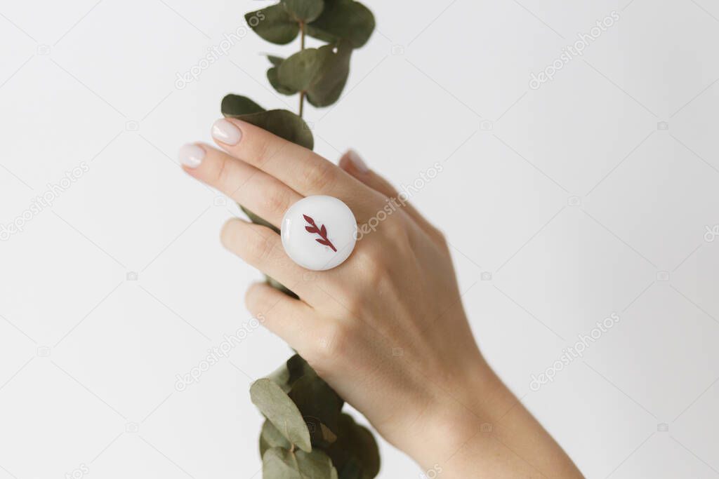 Stylish modern white round ring on beautiful hand with eucalyptus branch on white background. Unusual fashionable fused glass ring on female hand with white manicure. Eco and care concept